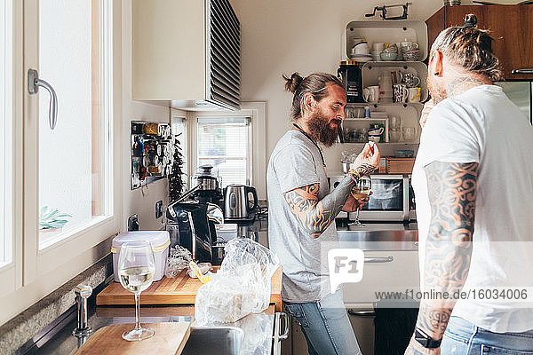 Two bearded tattooed men with long brunette hair standing in a kitchen  eating.