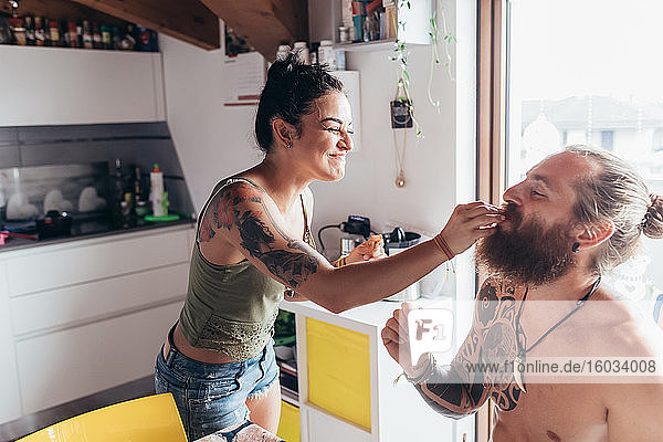 Bearded tattooed man with long brunette hair and woman with long brown hair in a kitchen  feeding each other.