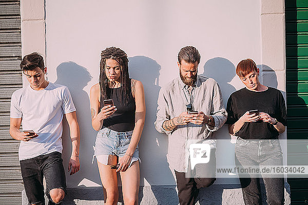 Two young women and men wearing casual clothes leaning against wall  using mobile phones.
