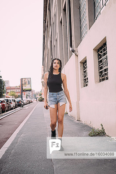 Portrait of young woman with dark brown hair  wearing black vest and denim shorts  walking down street.