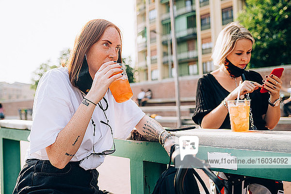 Zwei junge Frauen sitzen im Freien auf einer Bank  trinken und telefonieren.