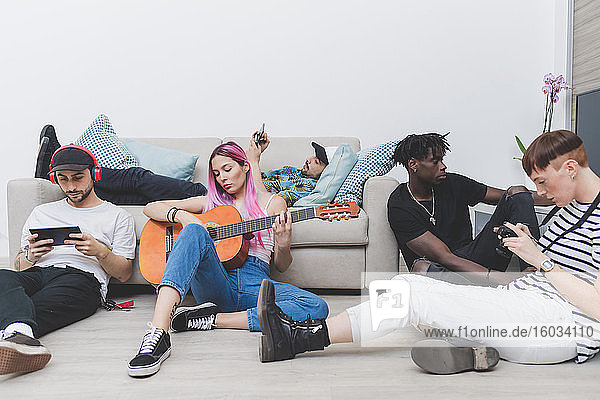 Group of young men and women sitting on living room floor  checking their mobile phones  woman with long pink hair playing guitar.