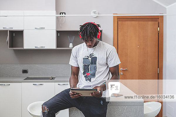 Young man with short dreadlocks sitting in a kitchen  wearing headphones and holding digital tablet.