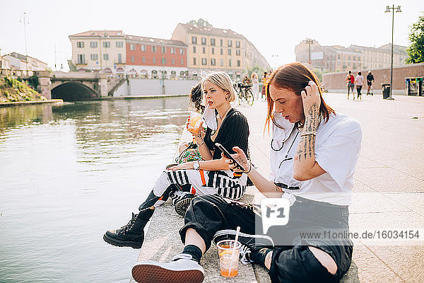 Drei junge Frauen sitzen an einem Flussufer  trinken und telefonieren.