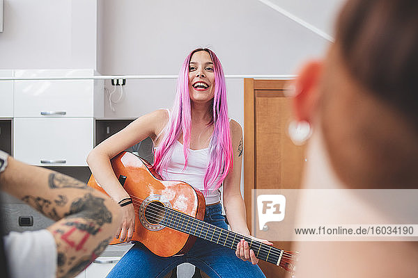 Young woman with long pink hair sitting in a kitchen  playing guitar.