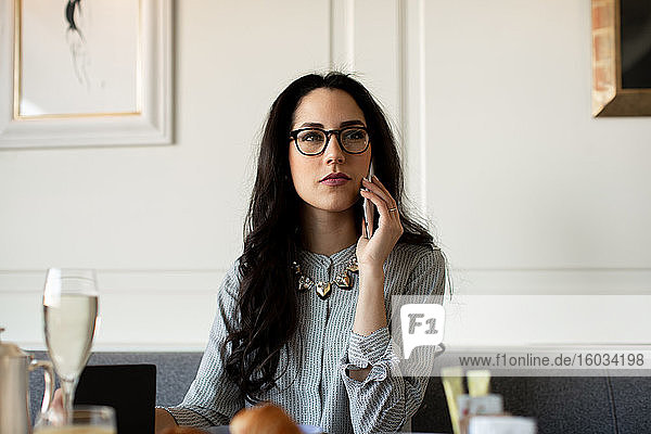 Woman with long black hair wearing glasses sitting at a restaurant table  using mobile phone.