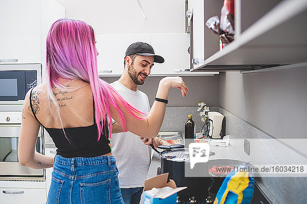 Junge Frau mit langen rosa Haaren und bärtigem Mann mit Baseballmütze steht in einer Küche und kocht.