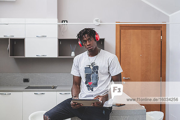 Young man with short dreadlocks sitting in a kitchen  wearing headphones and holding digital tablet.
