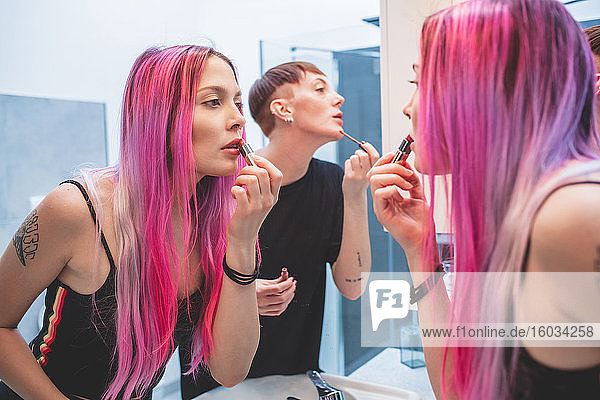 Young woman with long pink hair and woman with short red hair standing in front of mirror  applying lipstick.