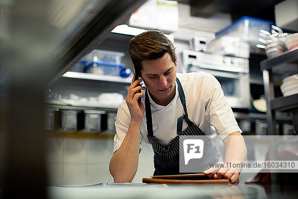 Junger männlicher Koch liest digitales Tablet und spricht über Smartphone in der Küche