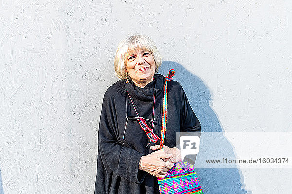 Porträt einer älteren Frau mit blonden Haaren in schwarzem Top  die vor einer weißen Wand steht und in die Kamera lächelt.