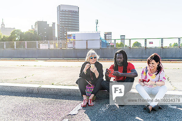 Schwarzer Mann mit Dreadlocks und zwei kaukasische Frauen sitzen auf dem Bürgersteig und überprüfen ihre Mobiltelefone.