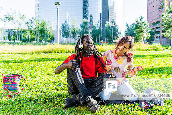 Schwarzer Mann mit Dreadlocks und kaukasische Frau sitzen auf dem Rasen und hören Musik.