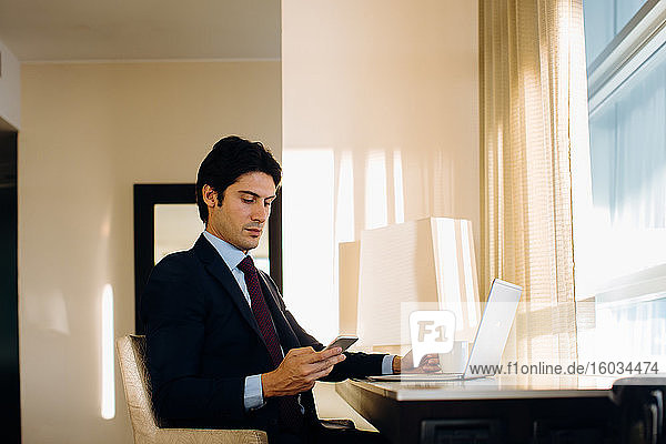 Businessman texting while using laptop by hotel room window