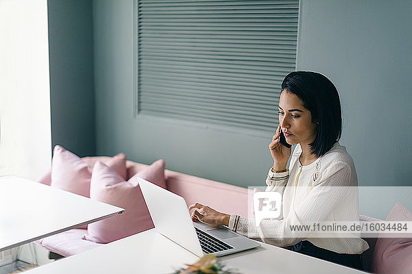 Young businesswoman at hotel table looking laptop and using smartphone