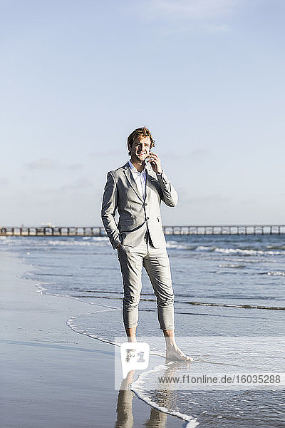 Barfüssiger Geschäftsmann spricht mit einem Smartphone am sonnigen Strand des Ozeans  Los Angeles  Kalifornien
