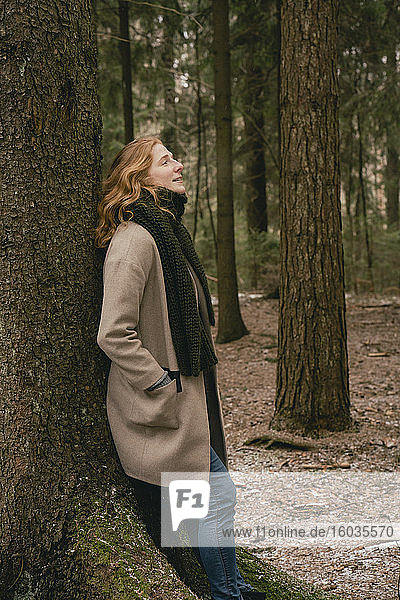 Gelassene rothaarige Frau in Wollmantel und Schal  die sich im Wald an einen Baum lehnt