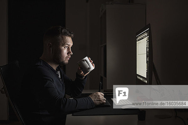 Mann mit Kaffee arbeitet spät am Computer in einem dunklen Raum