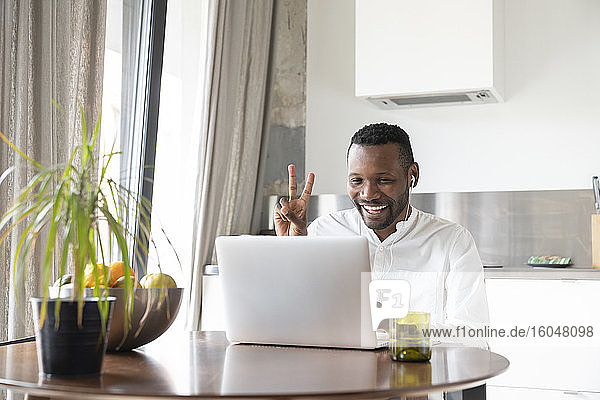 Porträt eines glücklichen Mannes  der während eines Videochats am Küchentisch sitzt und ein Siegeszeichen zeigt