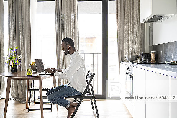 Lächelnder Mann sitzt am Tisch in einer modernen Wohnung und benutzt Laptop und Kopfhörer