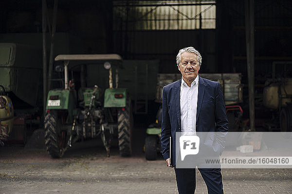 Porträt eines älteren Geschäftsmannes mit Laptop auf einem Bauernhof mit Traktor in einer Scheune