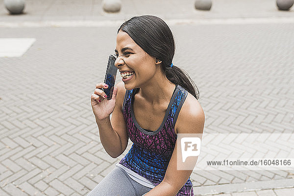 Fröhliche junge Frau  die mit ihrem Smartphone spricht  während sie auf dem Gehweg sitzt
