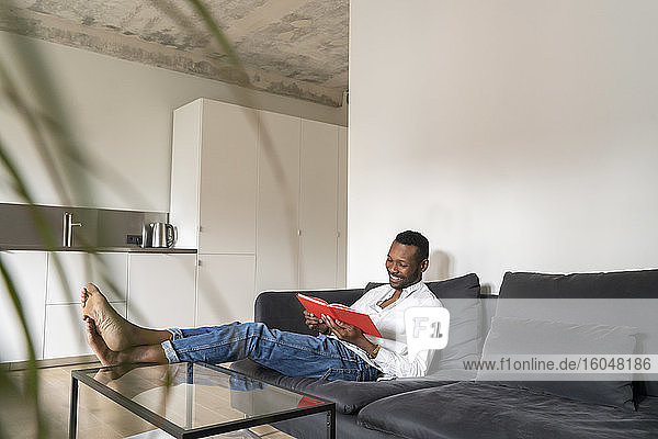 Lächelnder Mann sitzt auf der Couch in einer modernen Wohnung und liest ein Buch