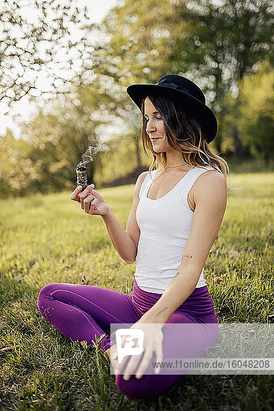 Smiling woman wearing hat holding incense while sitting on grassy land in park