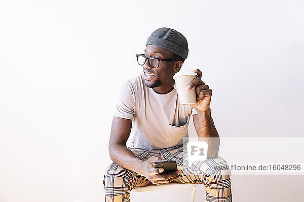 Junger Mann hält Handy und Kaffeetasse in der Hand und schaut weg vor weißem Hintergrund