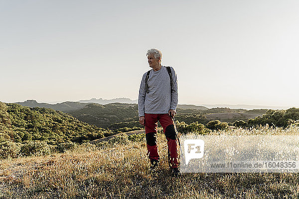 Senior male hiker looking away while standing with hiking pole on plants against clear sky