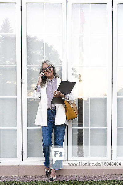 Lächelnde Unternehmerin  die mit einem Smartphone spricht und eine Akte gegen die Bürotür hält