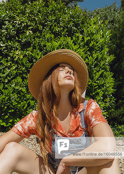 Schöne rothaarige Frau mit geschlossenen Augen sitzen gegen Pflanzen im Hinterhof auf sonnigen Tag