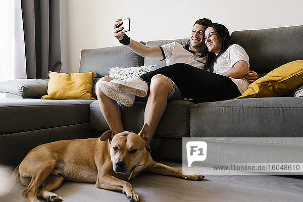 Paar macht Selfie auf Sofa in der Nähe von Hund zu Hause