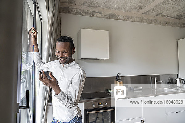 Porträt eines lächelnden Mannes  der in seiner Küche an der Balkontür steht und auf sein Mobiltelefon schaut