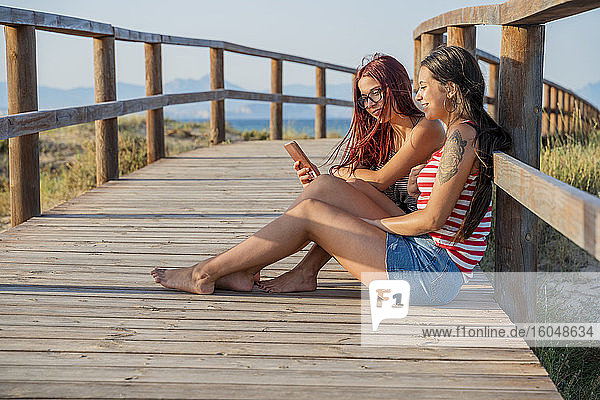 Teenage girls using smart phone while sitting on boardwalk at beach