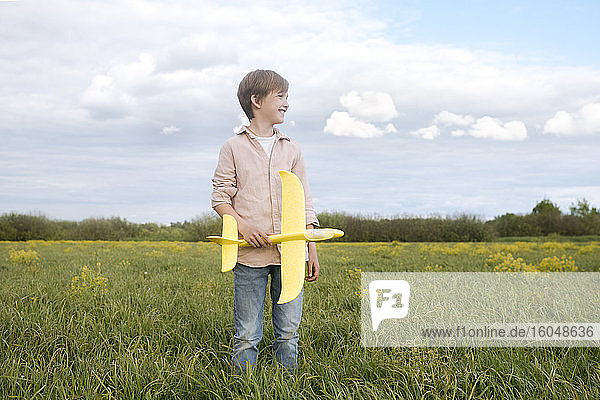 Lächelnder Junge  der ein Spielzeugflugzeug in der Hand hält und auf einem Rapsfeld in den Himmel schaut