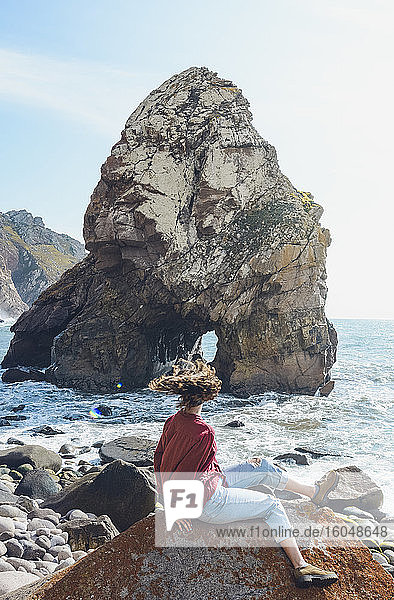 Sorglose Frau schüttelt den Kopf  während sie auf einem Felsen am Strand von Ursa sitzt  Portugal