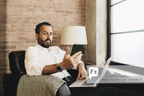 Mature man sitting in armchair  using laptop  holding smartphone
