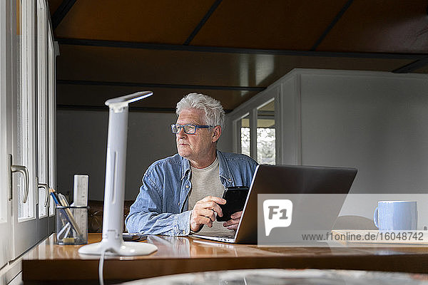 Thoughtful senior man holding smart phone with laptop on table at home