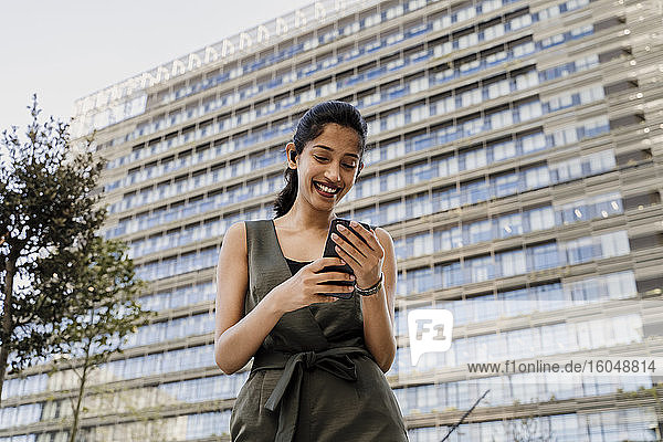 Lächelnde Geschäftsfrau  die ein Smartphone benutzt  während sie an einem Gebäude in der Stadt steht