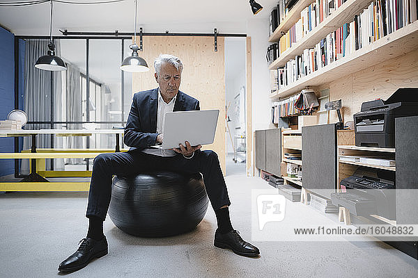 Älterer Geschäftsmann sitzt auf einem Fitnessball und benutzt einen Laptop