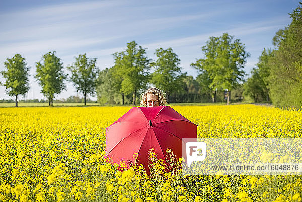 Ältere Frau mit rotem Regenschirm inmitten von Ölsaatenrapsen gegen den Himmel stehend