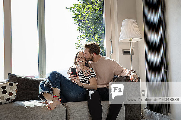 Mann küsst Frau beim Entspannen auf dem Sofa vor dem Fenster im Wohnzimmer