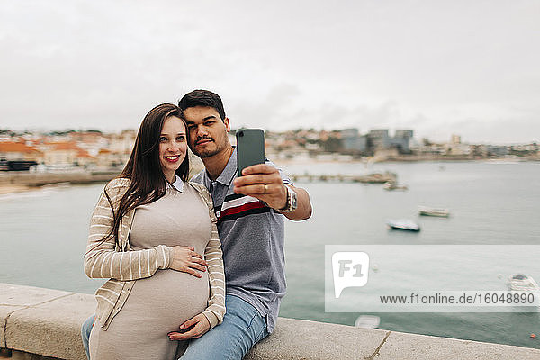 Expectant couple taking selfie through smart phone at harbor  Cascais  Portugal