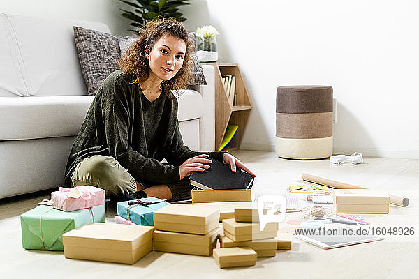 Porträt einer lächelnden jungen Frau  die zu Hause auf dem Boden sitzt und Geschenke einpackt