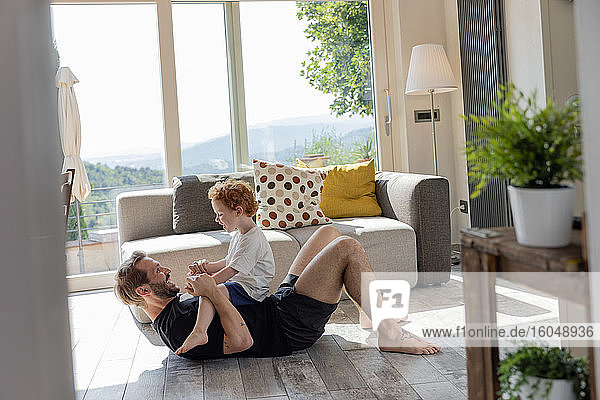 Happy man playing with cute son while lying by sofa in living room at home