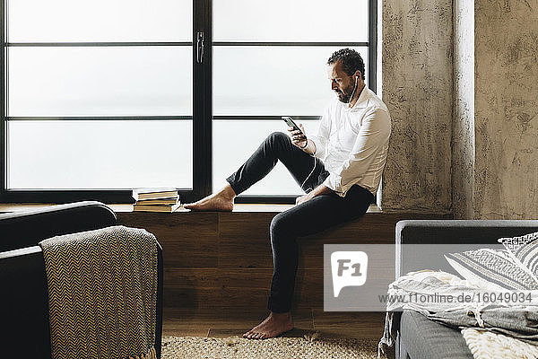 Mature man sitting barefoot on window sill  using smartphone and earphones