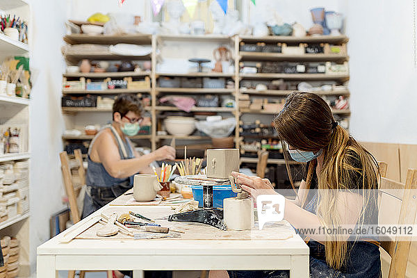Frauen mit Masken bei der Keramikherstellung auf einer Bank in einer Werkstatt