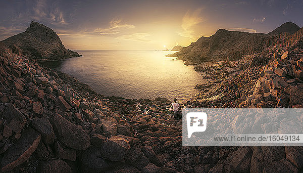 Spanien  Provinz Almeria  Frau sitzt allein am felsigen Küstenstrand im Naturpark Cabo de Gata-Nijar bei Sonnenuntergang