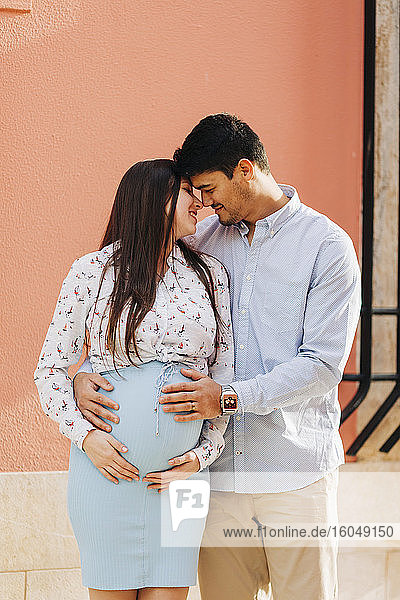 Romantisches schwangeres Paar  das mit den Händen auf dem Bauch vor einer rosa Wand in der Stadt steht
